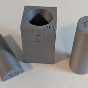 May include: Three gray 3D-printed objects: a square with a circular hole, a cylinder, and a half-cylinder. Each object has the number "9.0" or "0.6" printed on it. The objects are on a white surface.