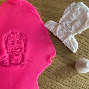Puede incluir: Un molde de plástico blanco de la cara de una persona con una expresión sonriente. El molde se está utilizando para crear una impresión en plastilina rosa.