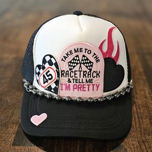 Puede incluir: Gorra de camionero negra y blanca con un parche rosa que dice "Take me to the racetrack & tell me I'm pretty". La gorra también tiene un corazón a cuadros con el número 45, un corazón rosa y una llama roja.