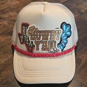Può includere: Un cappello da camionista beige con retro in rete. Il cappello presenta una toppa ricamata rosso, bianco e blu che dice "Howdy Y'all" e una toppa più piccola che dice "All Live In Tex". Il cappello ha anche un bordo di perline rosse a forma di stella.