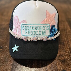 Puede incluir: Gorra de camionero negra con corona blanca y diseño de estrellas rosa y azul. La gorra tiene un lazo blanco y un parche azul y blanco que dice "Somebody's Problem". La gorra está decorada con una fila de cuentas brillantes.