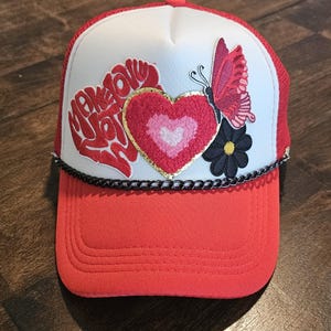 May include: A red and white trucker hat with a black chain trim. The hat features a red heart with a white heart inside, a butterfly, and a black flower with a yellow centre. The text "Make Love Not War" is embroidered on the hat.