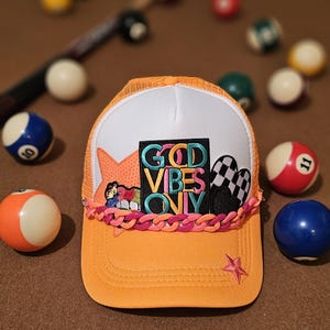 Puede incluir: Gorra de camionero naranja y blanca con parte trasera de malla. La gorra tiene un ribete de cadena rosa y naranja y un parche que dice "God Vibes Only".