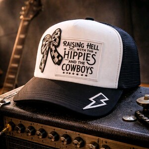 Puede incluir: Gorra de camionero en blanco y negro con un panel frontal blanco con un lazo a cuadros y el texto "Raising Hell with the Hippies and the Cowboys". Un rayo blanco está en el borde.