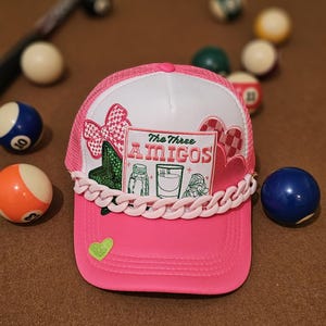 May include: Pink trucker hat with a white crown and a pink mesh back. The hat has a variety of colorful patches, including a green cactus, a pink heart, and a white patch with the text "The Three Amigos". The hat also has a pink chain around the brim.