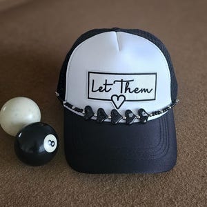 May include: A black and white trucker hat with the text "Let Them" and a heart graphic. The hat has a black brim and a beaded accent with heart-shaped charms. An 8-ball and a white ball are next to the hat.