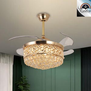 Puede incluir: Un ventilador de techo dorado con detalles de cristal y aspas transparentes. El ventilador está encendido y las aspas giran. El ventilador está en una habitación con paredes blancas y una pared verde.