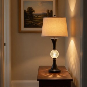Op de afbeelding: Een decoratieve tafellamp met een beige lampenkap en een zwarte voet. De lamp heeft een kristallen bol in het midden en staat op een donkere houten tafel. Een landschapsschilderij hangt op de achtergrond aan de muur.