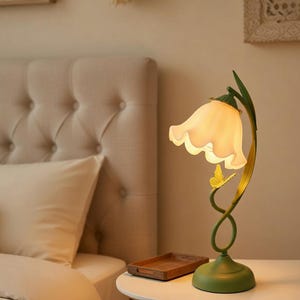 May include: A decorative table lamp in the shape of a flower. The lamp has a white, scalloped shade and a green base with a yellow butterfly accent. The lamp is on a white table next to a bed.
