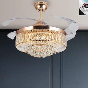 May include: A gold ceiling fan with a crystal chandelier design. The fan blades are white and the light fixture is surrounded by clear crystals.