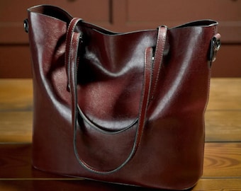 May include: A burgundy leather tote bag with two handles and a shoulder strap. The bag has a rectangular shape with a slightly curved top and a smooth, glossy finish. The bag is sitting on a wooden surface.