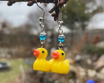 May include: A pair of silver earrings with yellow rubber duck charms and blue glass beads. The earrings are hanging from a branch.
