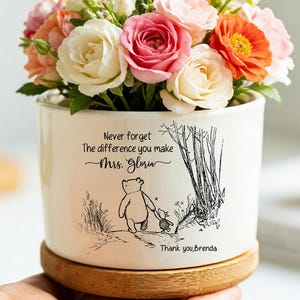 Può includere: Un vaso di fiori in ceramica bianca con base in legno, pieno di rose colorate. Il vaso presenta un'illustrazione di Winnie the Pooh e il testo "Never forget the difference you make Mrs. Gloria. Thank you Brenda."