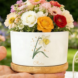 Puede incluir: Una maceta de cerámica blanca con base de bambú, llena de un colorido surtido de flores. La maceta presenta una ilustración de narciso y el nombre "Olivia". Las flores incluyen rosas, margaritas y otras flores.