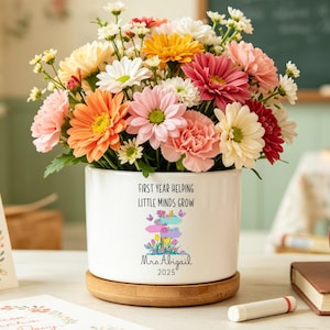 Puede incluir: Un vibrante ramo de flores coloridas en una maceta de cerámica blanca con el texto "FIRST YEAR HELPING LITTLE MINDS GROW". Una manzana roja está sobre un libro, y una tarjeta dice "American Tea Day".