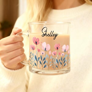 May include: A clear glass mug with a handle, filled with a light brown liquid. The mug is decorated with pink and light pink floral designs and blue leaves. The name "Shelley" is written in black script on the mug.