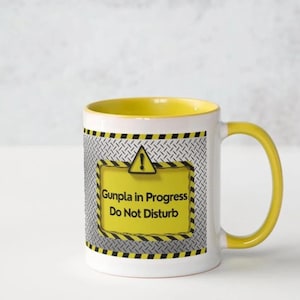 May include: White ceramic mug with a yellow interior and handle. The mug features a yellow warning sign with the text "Gunpla in Progress Do Not Disturb" on a silver diamond plate background.