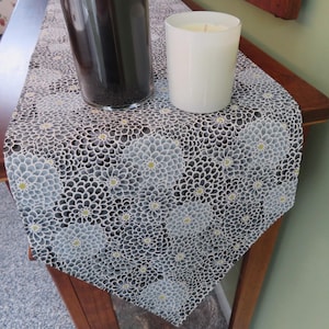 May include: A floral table runner with a repeating pattern of gray and white flowers with yellow centers on a black background. The runner is draped over a wooden table, with a glass vase and a white candle in a glass holder on top.