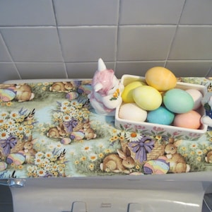 May include: A white toilet bowl with a floral patterned toilet seat cover. The cover features a basket filled with pastel coloured Easter eggs and two bunny figurines.