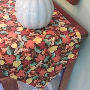 May include: A brown table runner with a fall leaf and pine cone pattern. The runner is decorated with a white pumpkin.