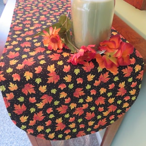 May include: A black table runner with a fall leaf pattern. The leaves are in shades of orange, yellow, and green. The runner is decorated with small gold dots.