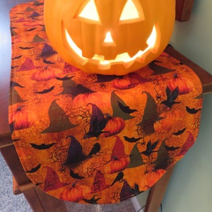 May include: Orange and black Halloween table runner with a witch hat and pumpkin pattern. The runner is made of fabric and has a stitched edge. A jack-o-lantern is sitting on top of the runner.