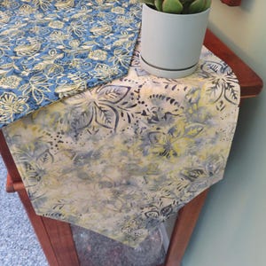 May include: A table runner featuring a dual-sided design with a blue fabric side displaying a dense yellow and white floral pattern and a cream-colored side with a muted gray and yellow batik-style floral print, topped with a small succulent.