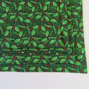 May include: A stack of folded green fabric with a repeating pattern of holly leaves and red berries. The fabric is a dark green color, and the holly leaves are a lighter shade of green. The berries are small and red.