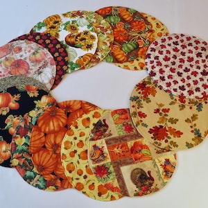 May include: A set of 10 round fabric placemats featuring a variety of autumnal designs, including pumpkins, leaves, turkeys, and acorns. The placemats are in shades of orange, brown, yellow, and red.