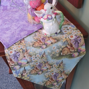 May include: A purple and blue table runner with a floral pattern featuring bunnies, baskets, and Easter eggs. The runner is decorated with a white ceramic bunny pitcher.