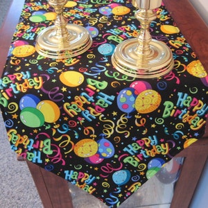 May include: A black table runner with a colorful "Happy Birthday" design. The runner features balloons in various colors, confetti, and the words "Happy Birthday" in a rainbow of colors. Two gold candle holders are on top of the runner.