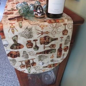 May include: A beige table runner with a wine-themed pattern featuring bottles, corkscrews, and grapes. The runner is draped over a wooden table, with wine glasses and a bottle of wine. The bottle has a white label with the year 2021.