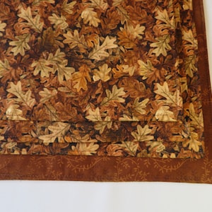 May include: A brown and beige fabric placemat with a pattern of autumn leaves. The placemat has a brown border with a vine pattern.