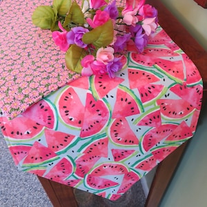 May include: Watermelon table runner with a white background and pink and green watercolor watermelon slices. The runner is decorated with a floral pattern on one end.
