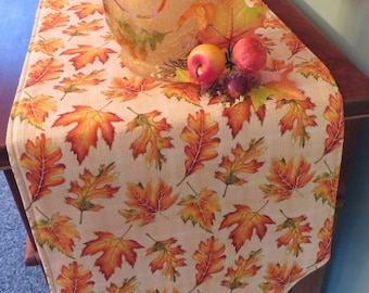 May include: A beige table runner with a pattern of orange and green autumn leaves. The runner is decorated with a glass vase and fall-themed decorations.