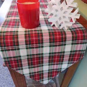 May include: Red, green, white, and pink plaid table runner with a white snowflake decoration and a red candle.