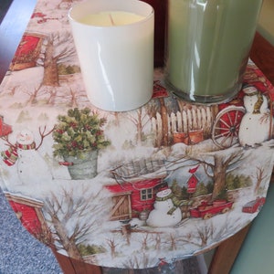May include: A white and red winter-themed table runner with a snowman, a red barn, and a Christmas tree. The runner is decorated with a white candle and a green candle in glass jars.