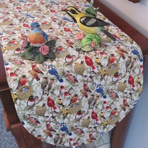 May include: A white table runner with a bird pattern. The runner features a variety of birds in different colors, including blue, red, yellow, and brown. The runner is decorated with a ceramic bird figurine.