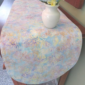 May include: A rectangular table runner featuring a soft watercolor-style floral pattern in shades of light blue, pale yellow, and lavender. The fabric has rounded ends and is topped with a small white ceramic vase containing yellow flowers.