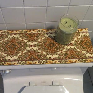 May include: A decorative toilet seat cover with a cream-colored background and an ornate pattern in shades of brown, green, and gold. A glass candle holder with a green candle sits on top of the cover.