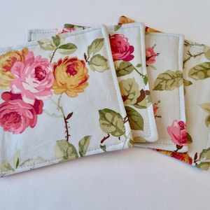 May include: Set of four square fabric coasters. Each coaster features a light blue background with a floral pattern of pink, yellow, and orange roses, along with green leaves and stems. The edges are finished with a white stitch.