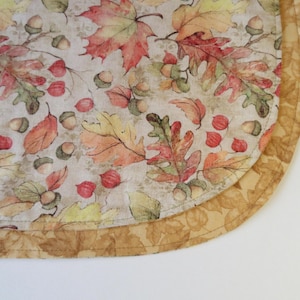 May include: A round fabric placemat with a beige background and a pattern of autumn leaves in shades of orange, yellow, and brown. The placemat has a double border of a lighter beige fabric.