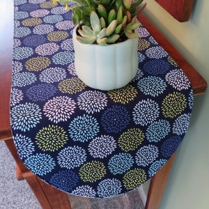 May include: A navy blue table runner with a white background and a pattern of green, blue, and white stylized flowers. The runner is on a wooden table with a potted succulent plant in the center.