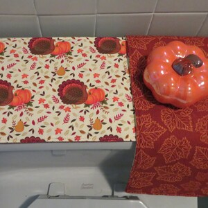 May include: A toilet seat cover and a decorative accent piece with an autumnal theme. The seat cover displays a repeating pattern of turkeys, pumpkins, and leaves on a cream background. A ceramic pumpkin sits on a burgundy leaf-patterned runner.