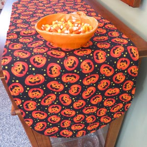 May include: A black table runner with a repeating pattern of orange jack-o'-lantern faces and yellow dots. A bowl filled with candy corn sits on the runner.