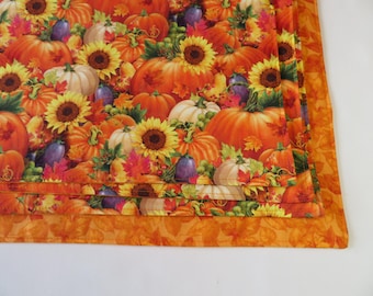 May include: A fabric placemat featuring a vibrant autumn pattern of orange pumpkins, yellow sunflowers, purple grapes, and red maple leaves. The placemat has a solid orange border with a subtle tonal leaf print and stitched edges.