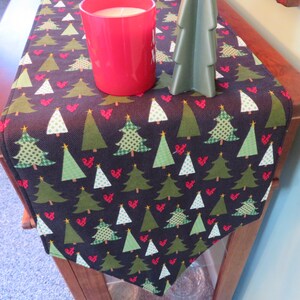 May include: A Christmas-themed table runner with a dark background featuring green and white Christmas trees and red hearts. A red candle holder and a green tree-shaped decoration sit on the runner, adding to the festive decor.