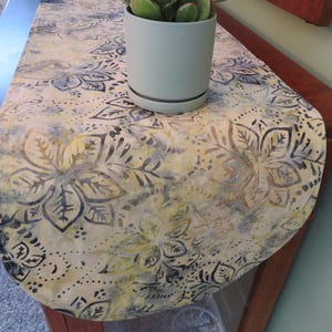 May include: A table runner featuring a batik-style floral pattern in shades of yellow, gray, and cream. The fabric has a rounded end and is decorated with dark gray outlines of flowers and leaves. A light gray cylindrical plant pot sits on top.