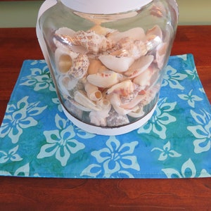 May include: A glass jar filled with seashells sits on a blue and green floral table runner. The runner has a white border and is made of fabric.