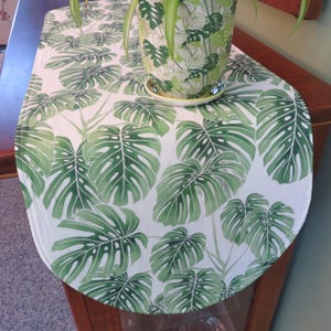 May include: A white table runner with a repeating pattern of large green monstera leaves. A matching plant pot with a green leaf design sits on the runner. The runner is placed on a wooden cabinet.